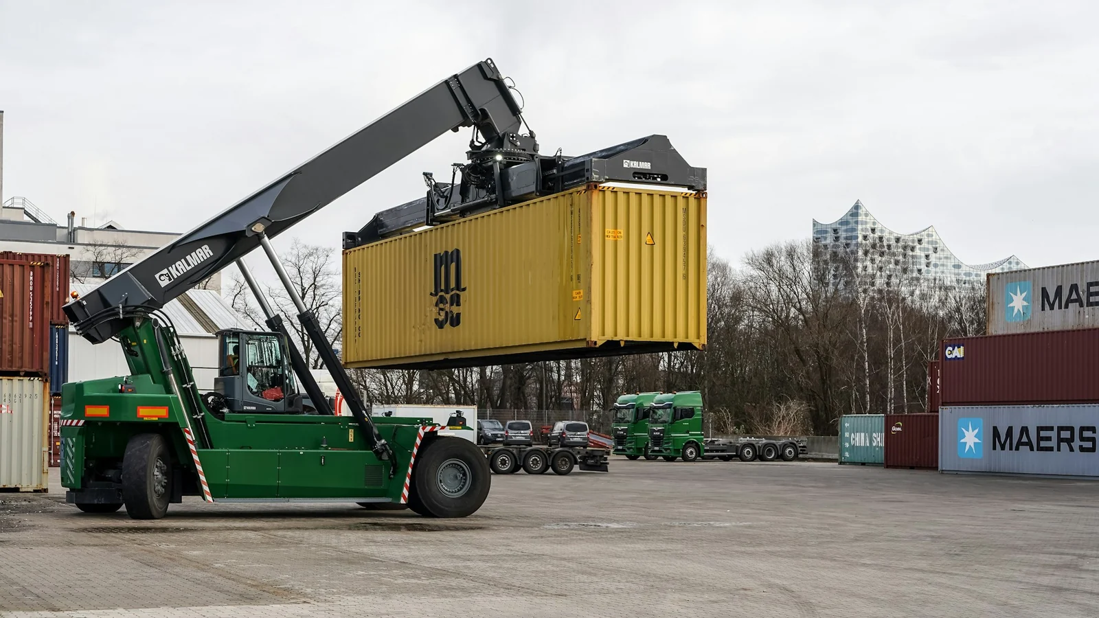 A green reach stacker lifting a yellow ocean shipping container in a container yard, with day-cab tractors and stacked containers in the background.